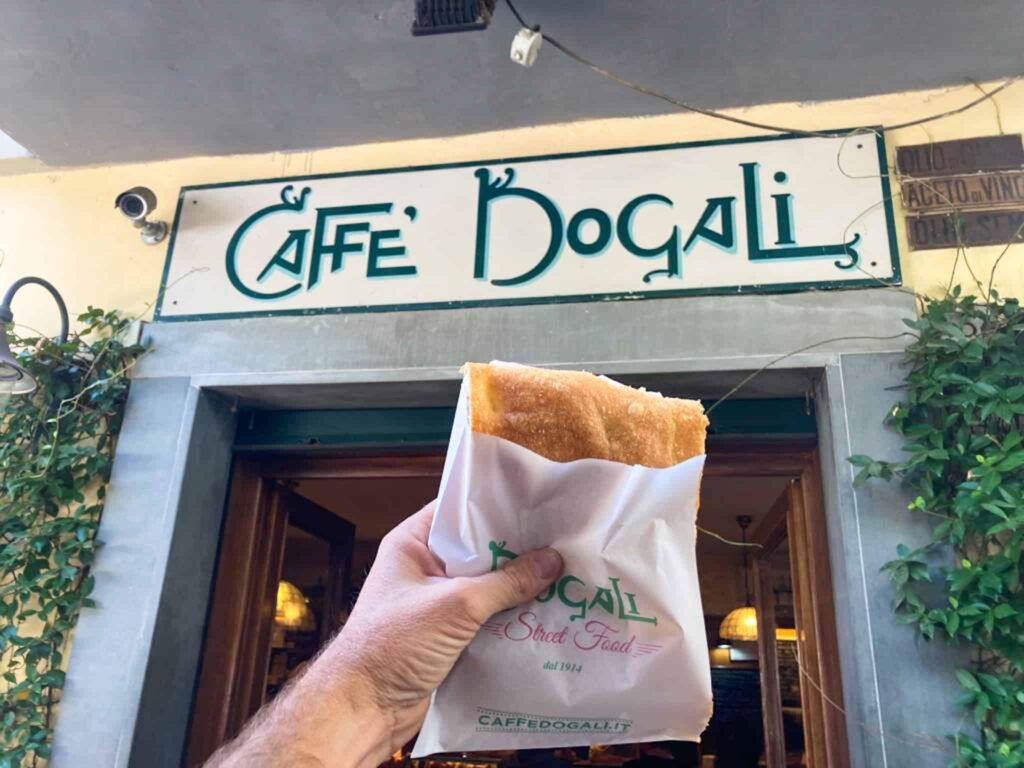 Una schiacciata nel sacchetto del Caffè Dogali, tenuta in mano davanti all'insegna del locale in viale Malta a Firenze