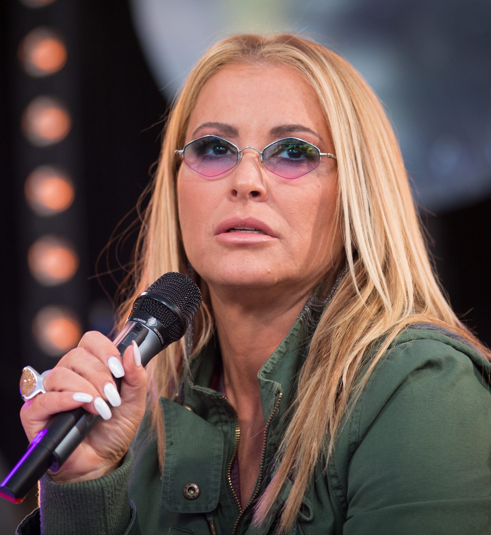 Anastacia in close-up, wearing purple-lensed glasses, with long blond hair and a microphone in her hand