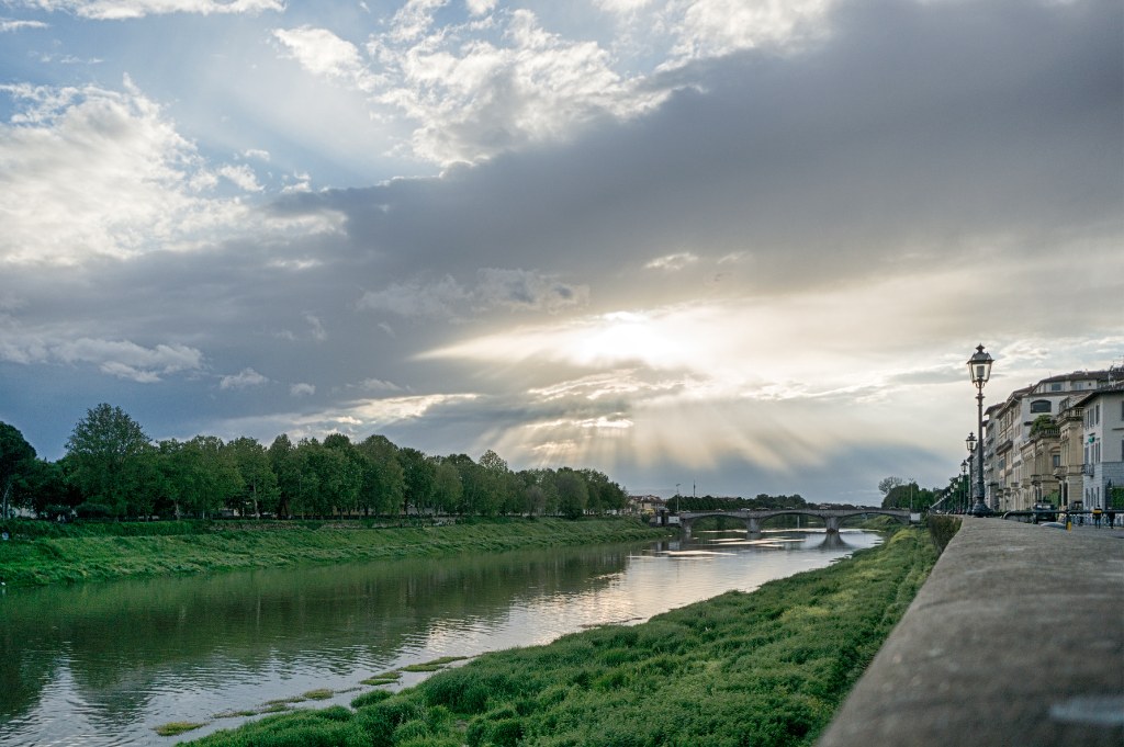 L'Arno fra due rive verdi sotto un cielo di nuvole attraversato dai raggi del sole, con un lampione sul lungarno
