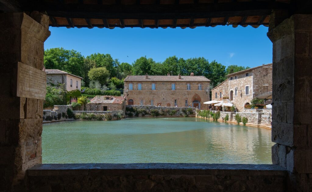 La grande vasca termale di Bagno Vignoni vista da un'apertura in pietra, con gli edifici del borgo sullo sfondo