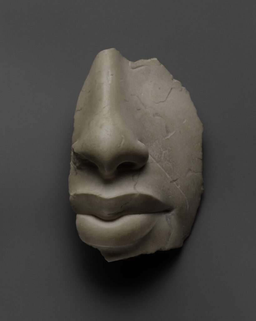 Frammento di volto in calcare chiaro con il naso e le labbra del faraone Akhenaton, fotografato su un fondo grigio uniforme