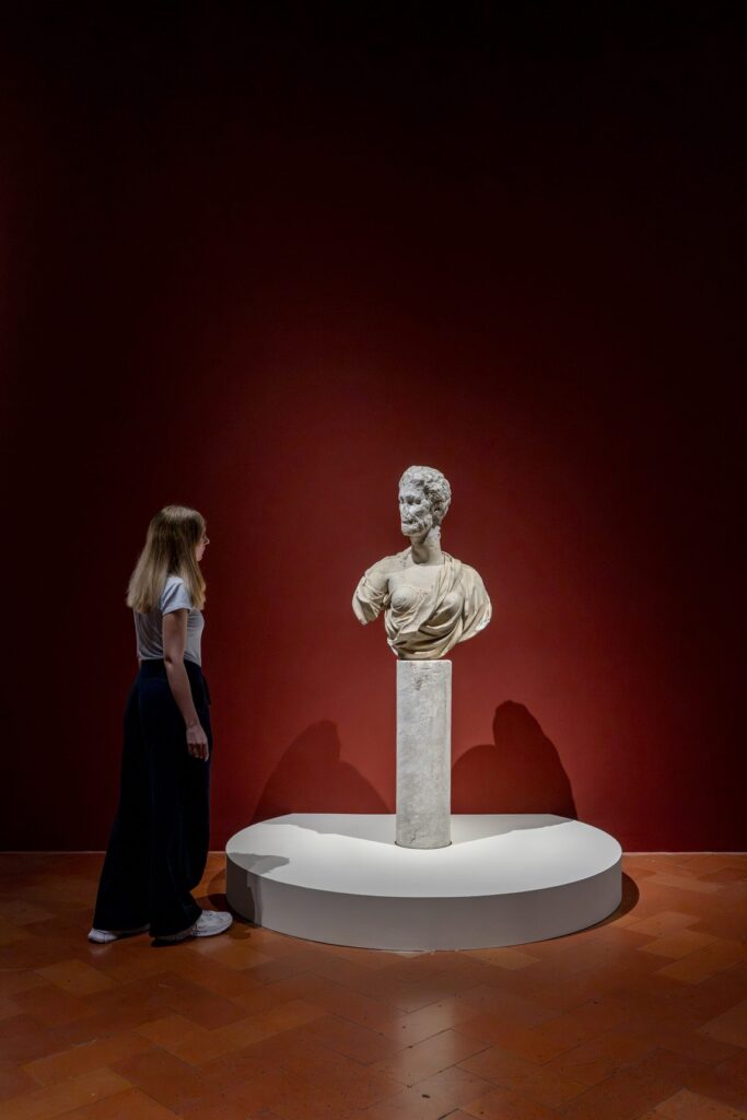 Una visitatrice osserva un busto in marmo su un piedistallo davanti a una parete rossa, nella mostra Broken a Palazzo Strozzi