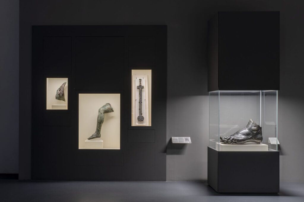 Frammenti in bronzo nella mostra Broken: una gamba e altri pezzi in nicchie illuminate su parete scura, un piede in vetrina