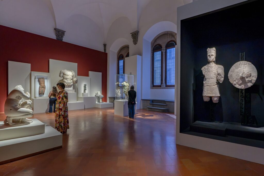 Una sala di Broken a Palazzo Strozzi: frammenti di statue su pedane bianche, parete rossa e un guerriero con scudo in nicchia