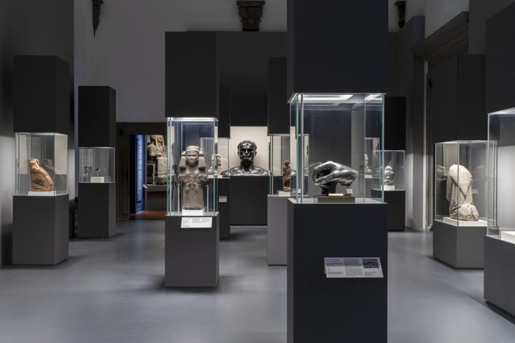 Una sala in penombra di Broken a Palazzo Strozzi: teste e frammenti di sculture dentro vetrine illuminate su basamenti grigi