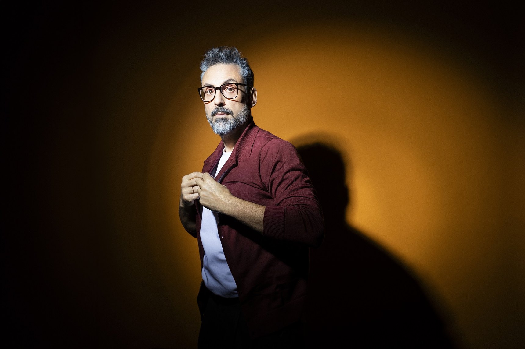 Brunori Sas in a burgundy jacket and glasses, lit by a warm light that casts his shadow on the wall