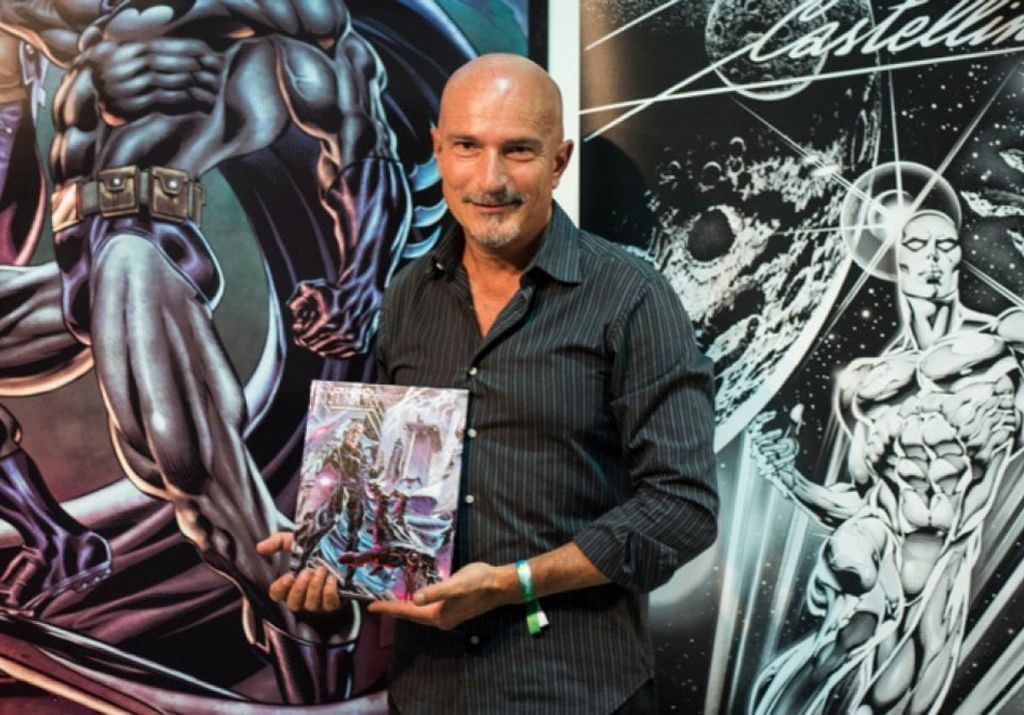 Claudio Castellini mostra una tavola a colori davanti ai suoi disegni di figure muscolose in bianco e nero