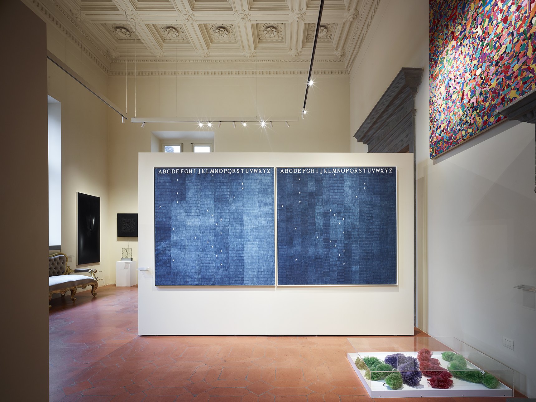 Sala con due grandi tele blu su una parete bianca, un'opera multicolore in alto e un'installazione colorata a terra