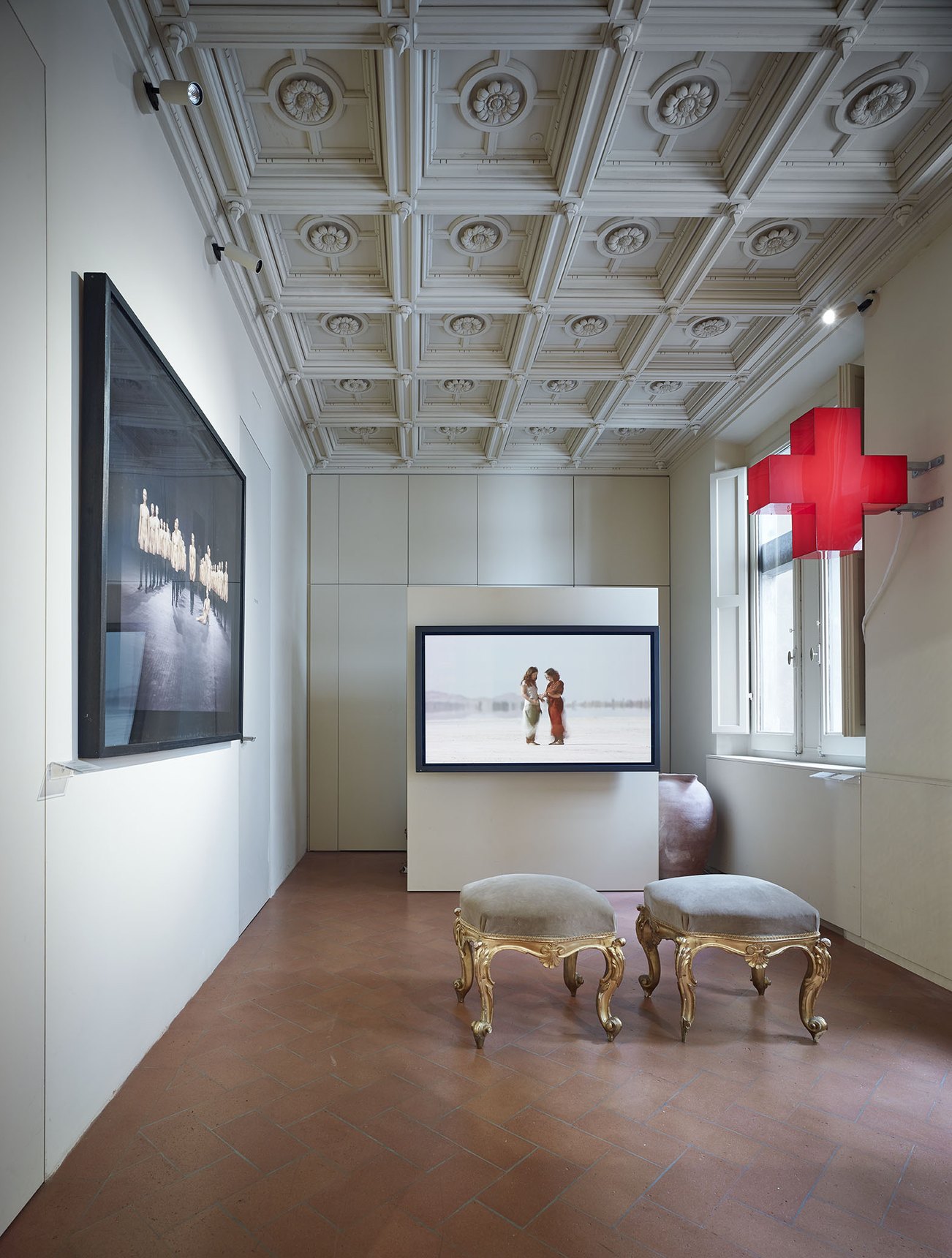 Sala dal soffitto a cassettoni con una croce rossa luminosa alla finestra, uno schermo con due figure e due sgabelli dorati