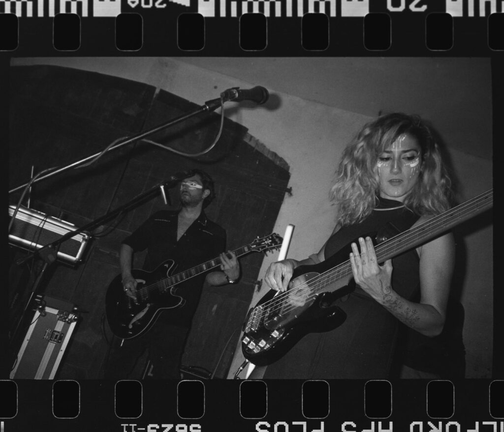 Black-and-white film shot: a curly-haired bassist and a guitarist playing beneath an arched vault