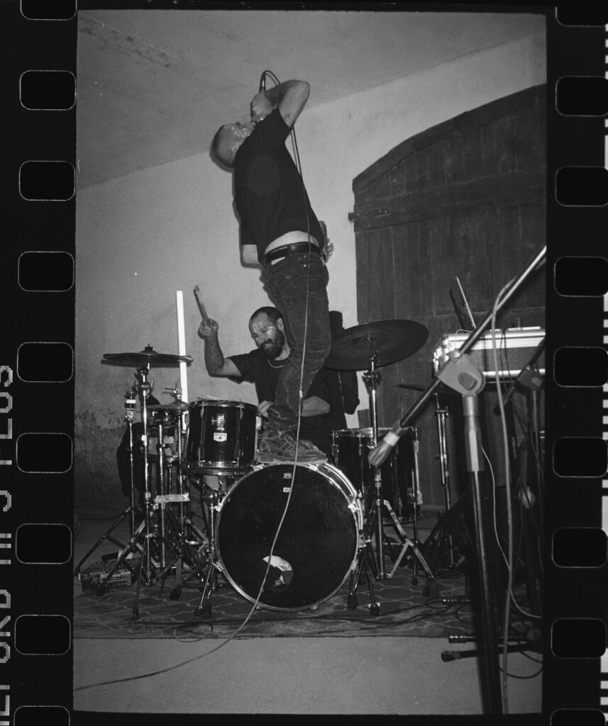 Black-and-white film shot: the singer arches backward while the drummer plays, in front of a large doorway