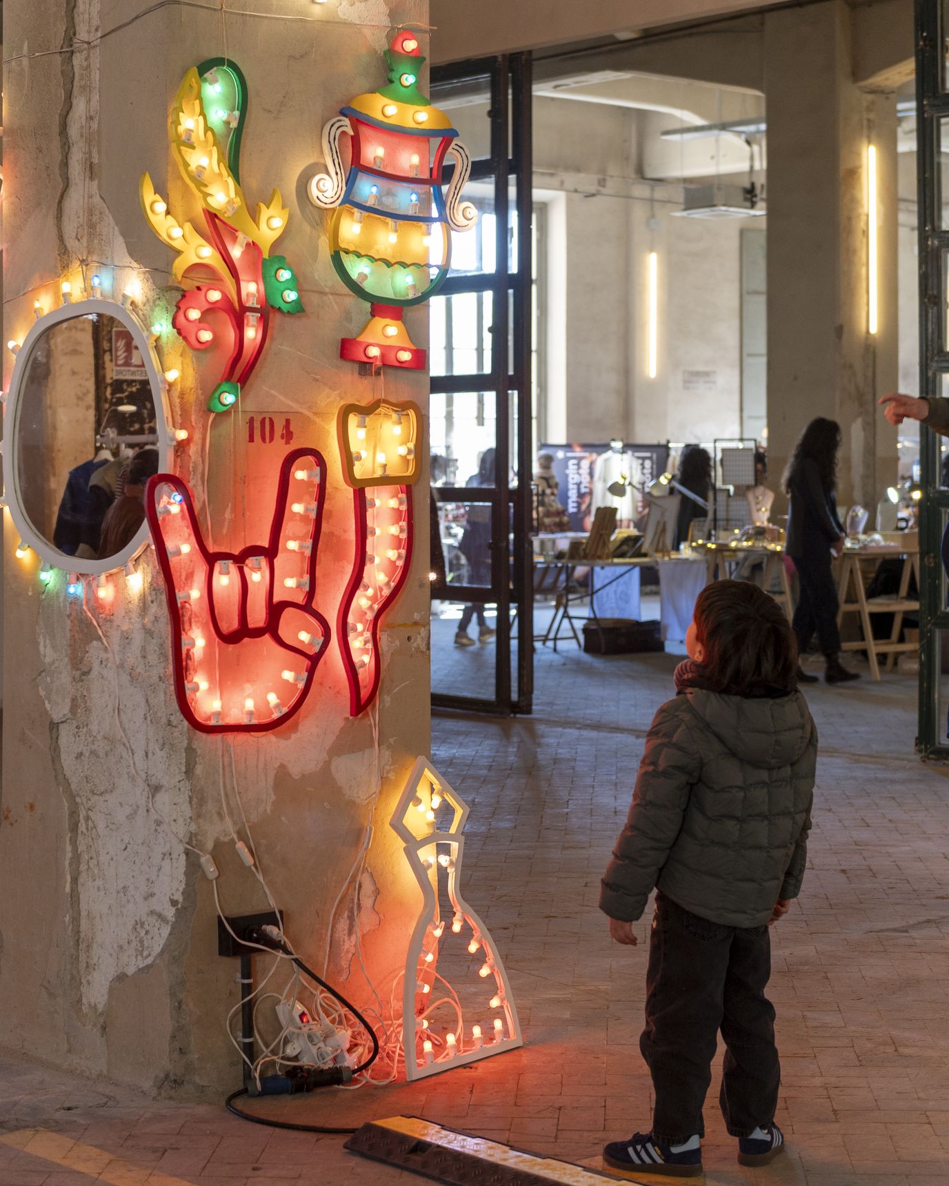 Un bambino guarda un’installazione luminosa al neon a forma di mano che fa il gesto I love you