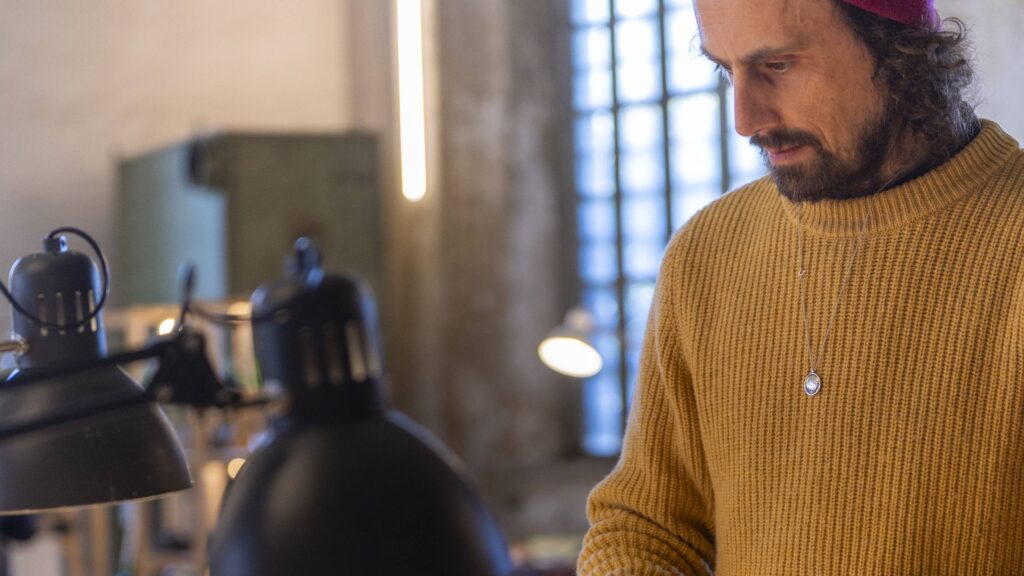 Artigiano con bandana osserva un gioiello dietro il banco, fra lampade da lavoro e finestre industriali