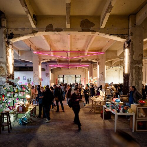 Il capannone industriale con i banchi del market, la folla di visitatori e i neon rosa sospesi in alto