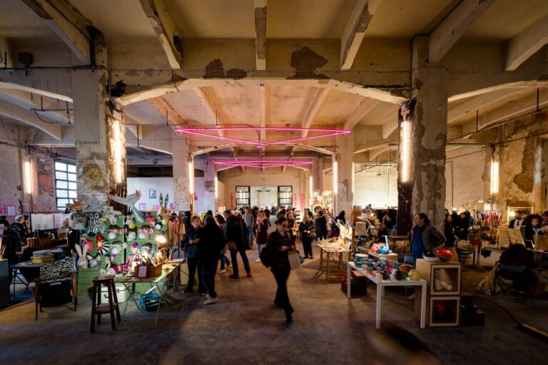 Il capannone industriale con i banchi del market, la folla di visitatori e i neon rosa sospesi in alto