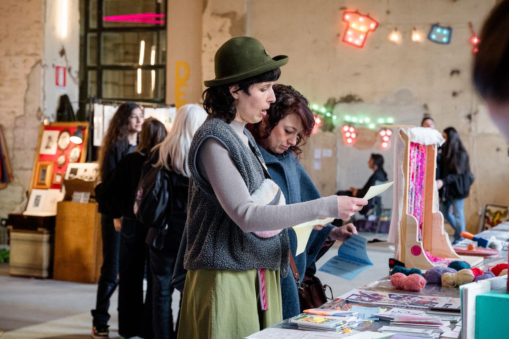 Visitatori davanti a un banco coperto di stampe e illustrazioni colorate, con luci al neon sullo sfondo