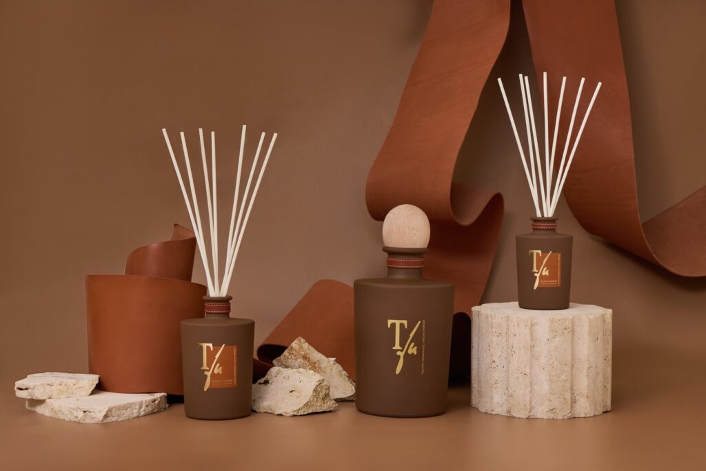 Reed diffusers and candles in leather-colored jars next to pale stones and a drape of leather, on a beige background
