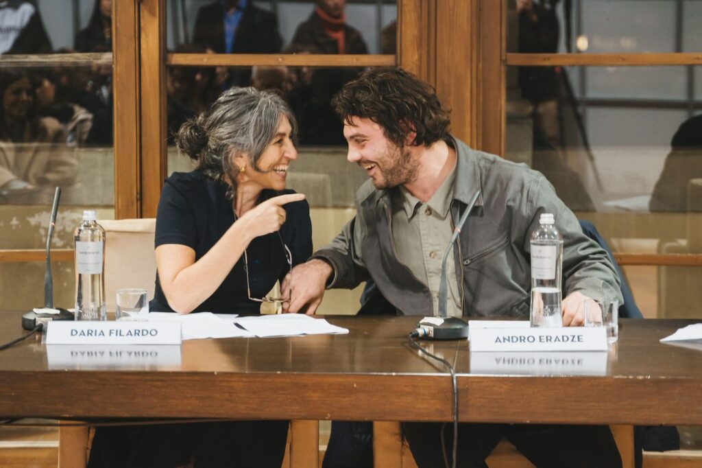 Daria Filardo e l'artista Andro Eradze seduti al tavolo di un incontro pubblico, lei lo indica sorridendo