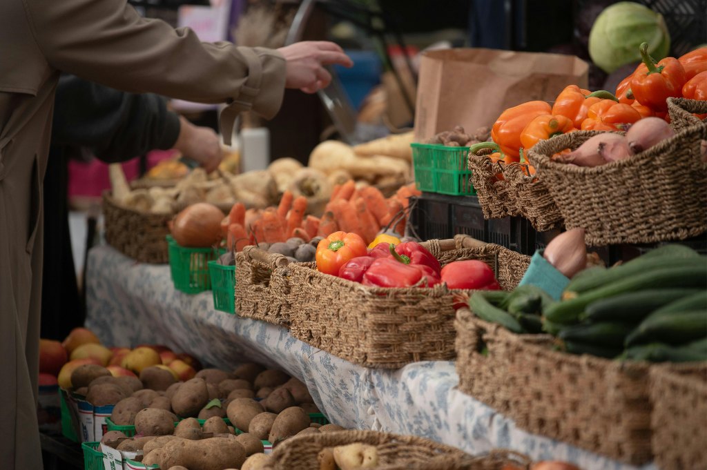 Wicker baskets of peppers, squash, zucchini and potatoes on a stall, with the hands of someone choosing
