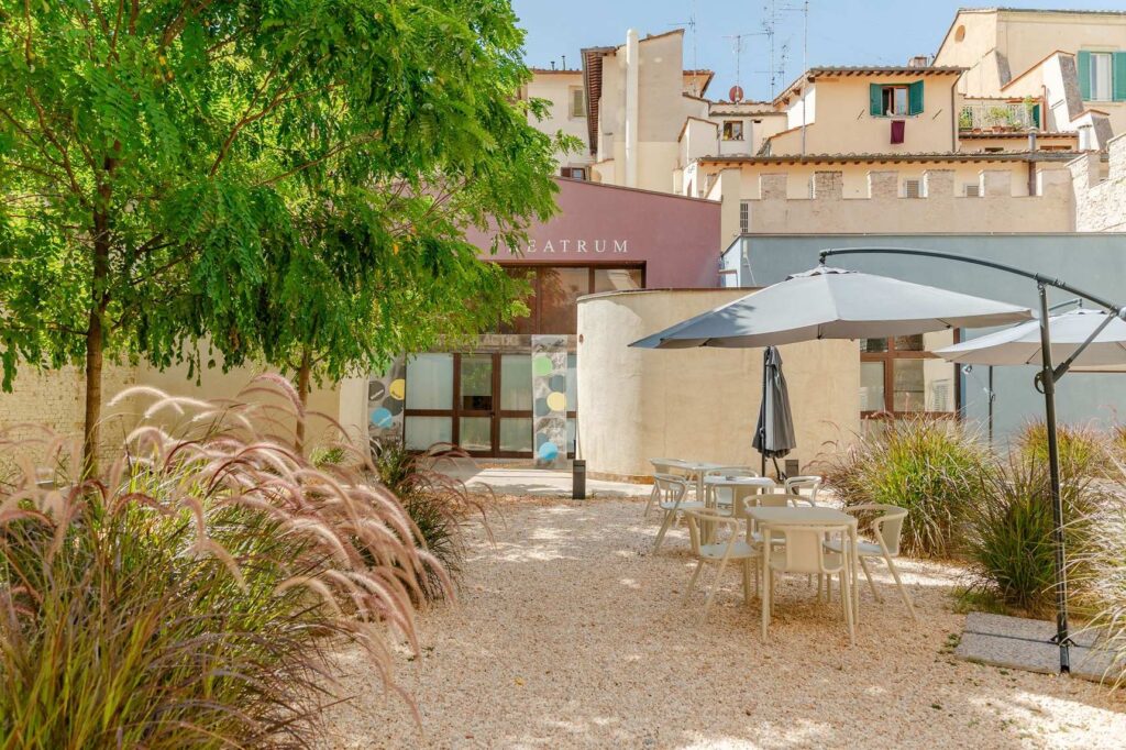 Il cortile dell'ex Teatro dell'Oriuolo a Firenze, con graminacee, un albero, ombrelloni e tavolini fra le case