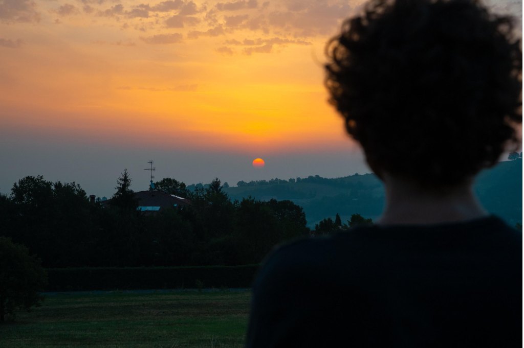 Un ragazzo dai capelli ricci ripreso di spalle, in controluce, guarda il sole che tramonta dietro le colline