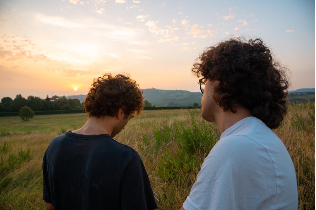 Due ragazzi ripresi di spalle in un prato d'erba alta guardano verso le colline mentre il sole tramonta all'orizzonte