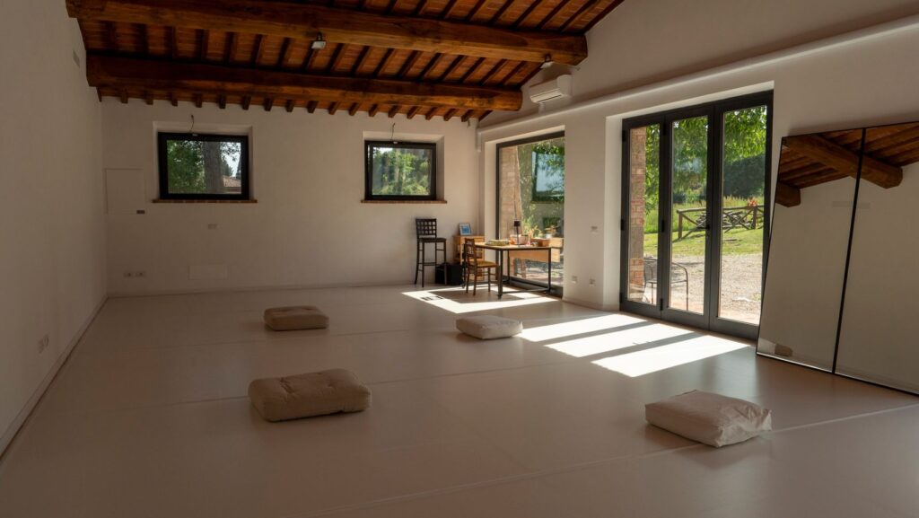 Uno studio vuoto di La CAP Re|Hub: pavimento bianco, cuscini a terra, travi di legno e vetrate aperte sul verde
