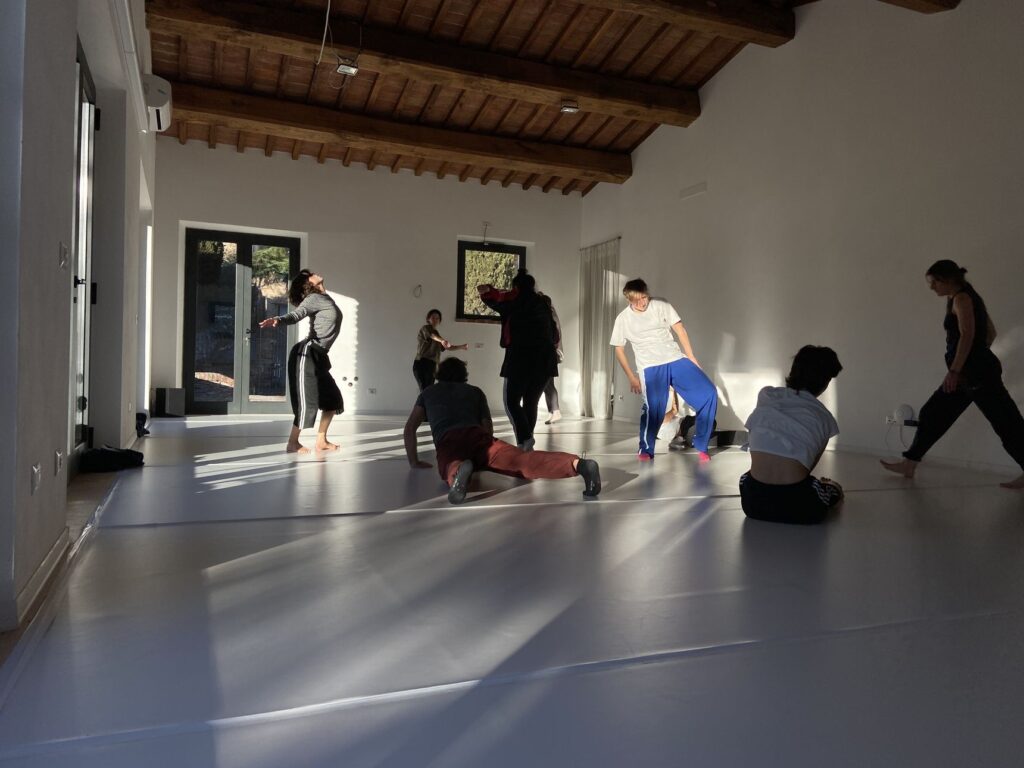 Residenti al lavoro nello studio di La CAP: corpi in movimento sul pavimento bianco, sotto il soffitto a travi di legno