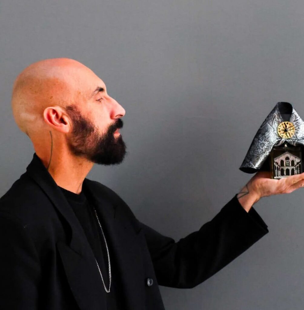 A man dressed in black, in profile, looks at a dark house-shaped object he holds in the palm of his hand
