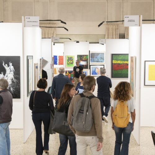 Visitatori camminano nel corridoio fra gli stand bianchi di Lucca Art Fair, con le opere appese ai lati