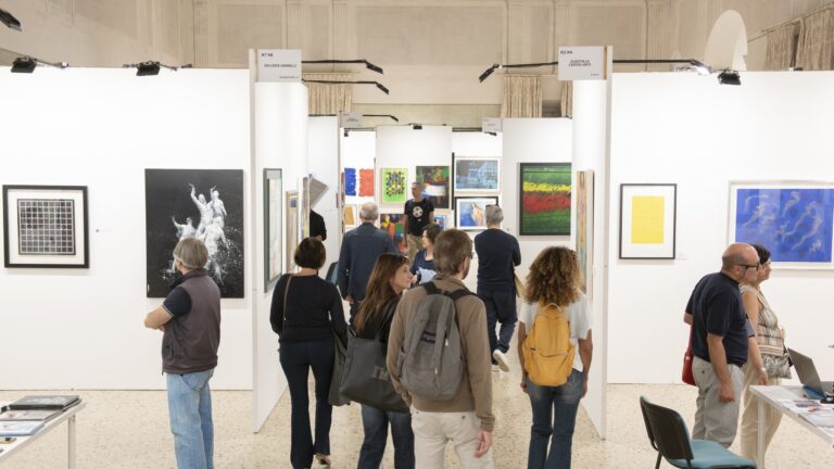 Visitatori camminano nel corridoio fra gli stand bianchi di Lucca Art Fair, con le opere appese ai lati