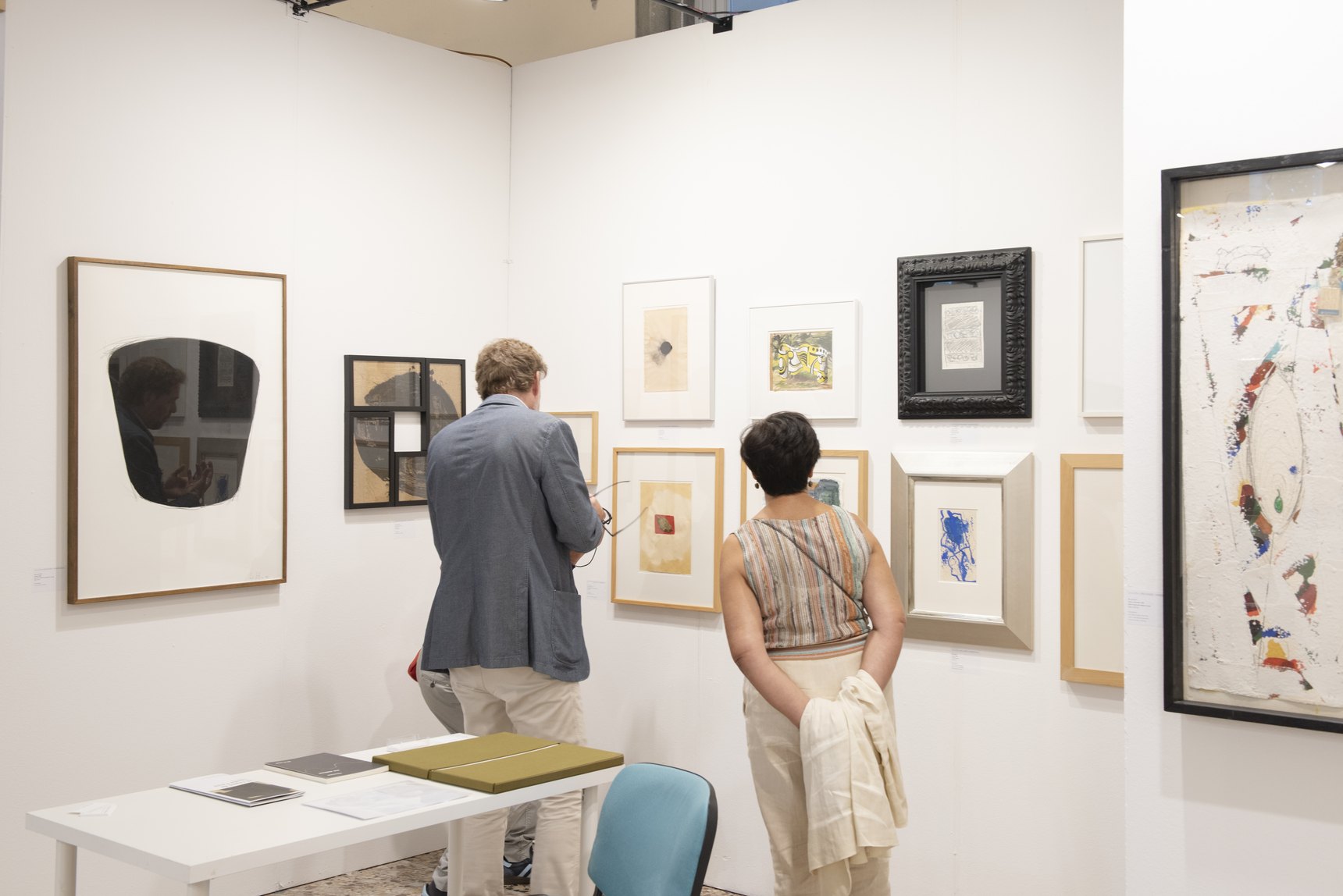 Due visitatori di spalle davanti a una parete di opere incorniciate in uno stand della fiera