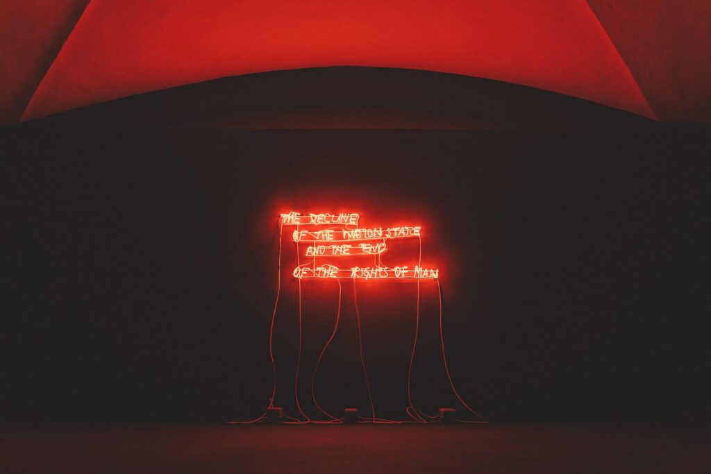 Una scritta al neon rosso accesa nel buio di una sala della mostra Alter Eva, sotto una volta illuminata di rosso