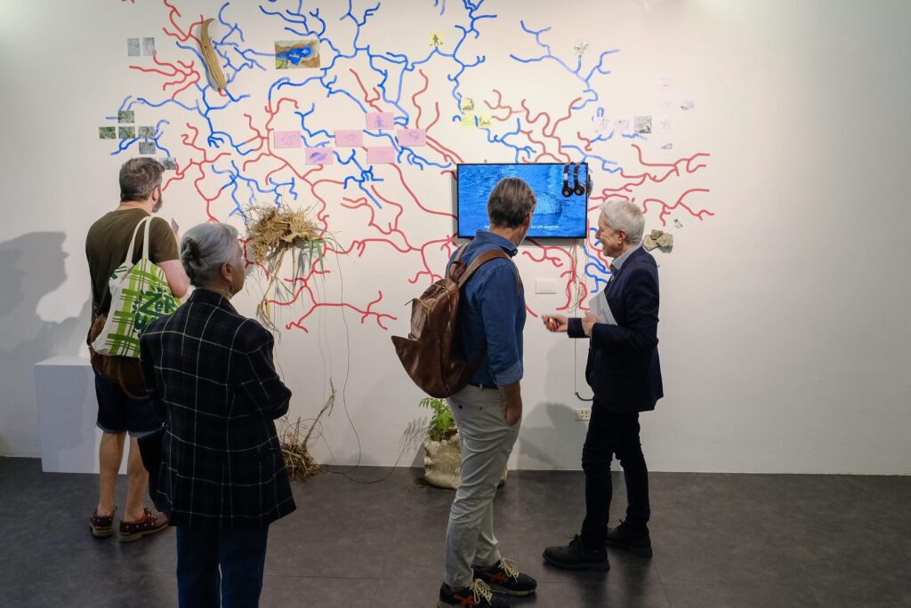Visitatori davanti a una parete coperta da un grande disegno di linee rosse e blu nella mostra Eco-Esistenze