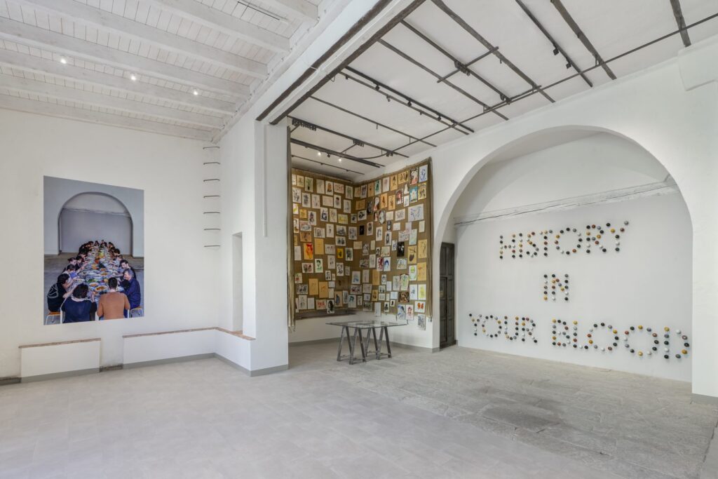 Veduta della mostra Teatro delle Relazioni: la scritta History in your blood su un arco bianco e una parete di immagini