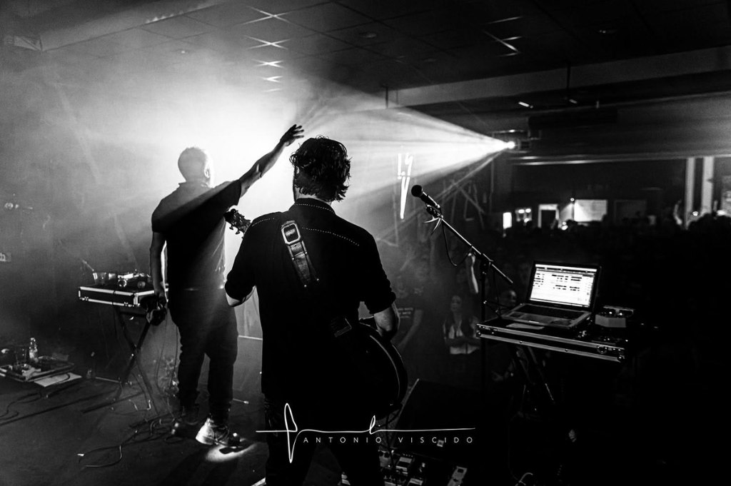 Black-and-white photo from the back of the stage: two musicians seen from behind, one raising an arm toward the crowd