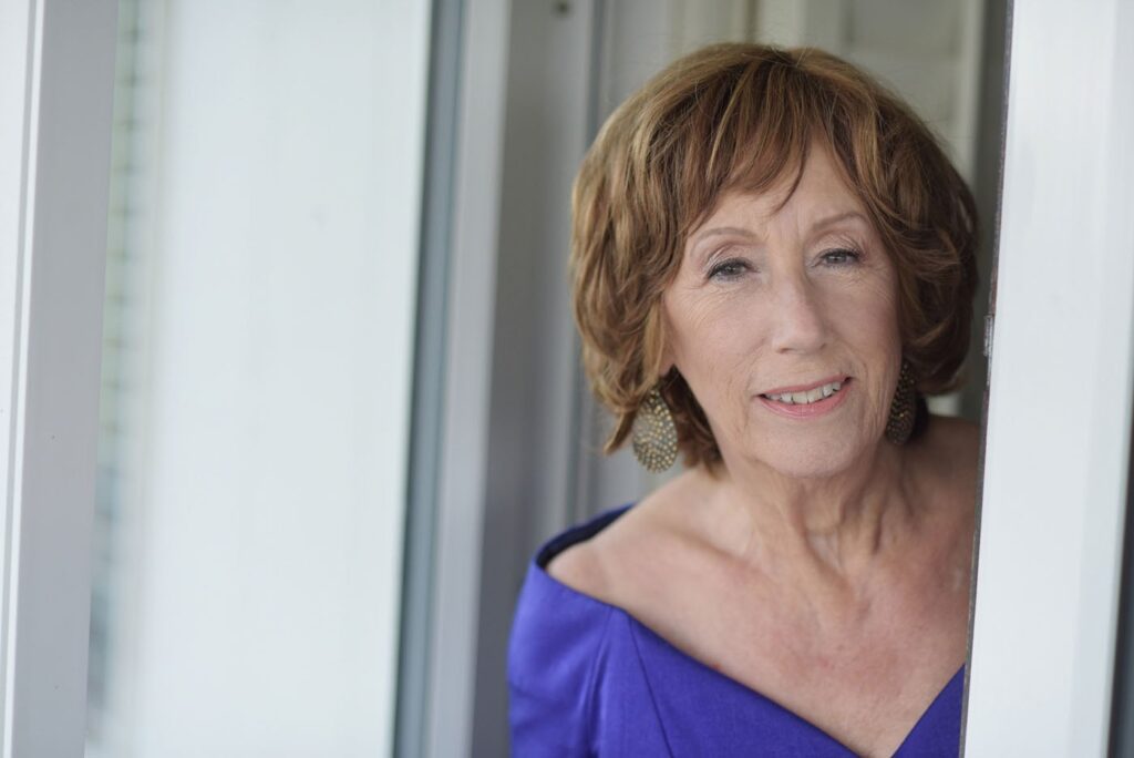 Ritratto della cantante Norma Winstone, capelli biondi corti e abito viola, davanti a una tenda chiara