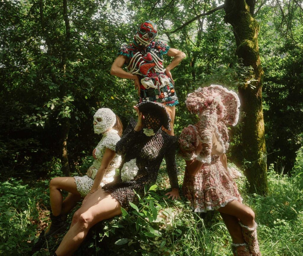Costumed figures among the trees of a forest, one with its face hidden behind a white skull-shaped mask
