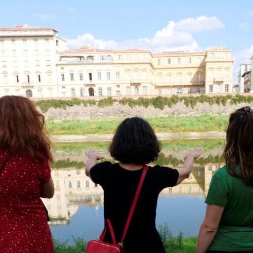 Tre donne di spalle sull'argine erboso dell'Arno guardano il fiume e i palazzi del lungarno riflessi nell'acqua