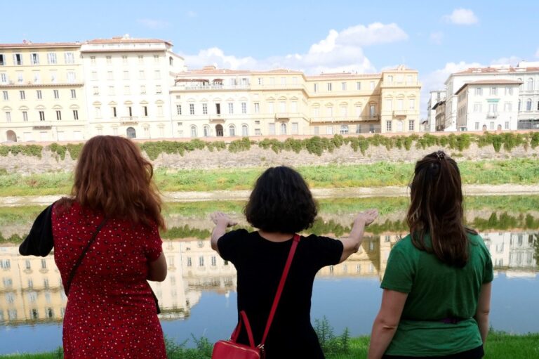 Tre donne di spalle sull'argine erboso dell'Arno guardano il fiume e i palazzi del lungarno riflessi nell'acqua
