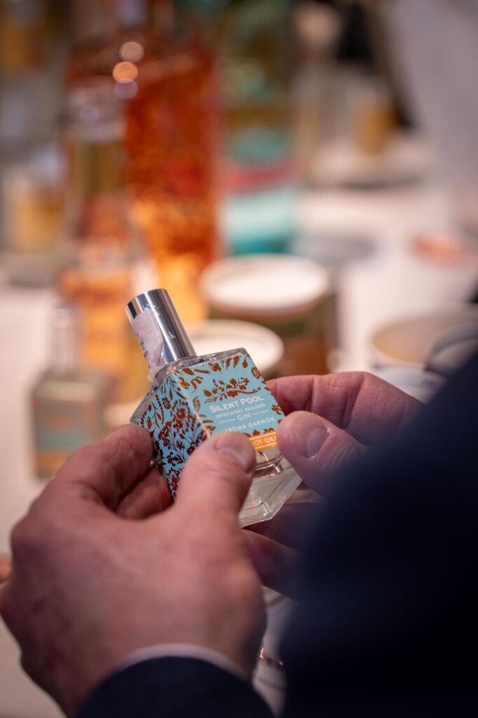 Two hands holding a small bottle with an ornate light-blue label, with other bottles blurred in the background