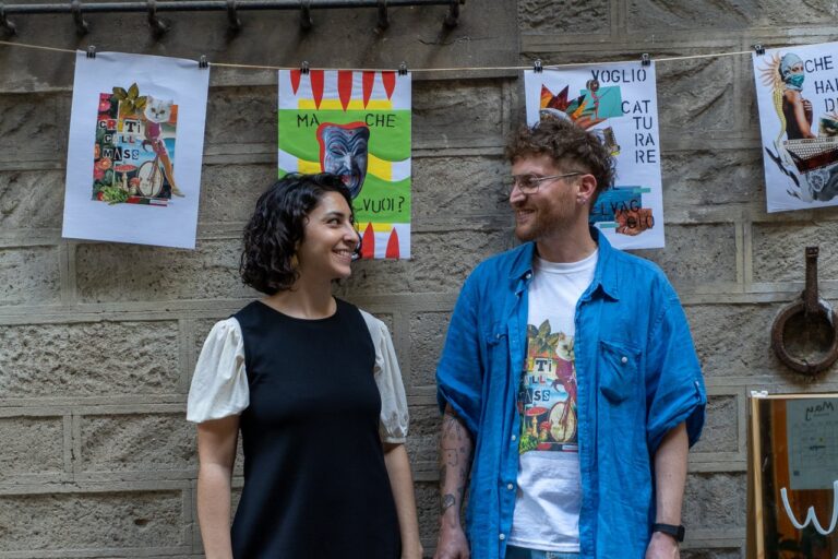 Due persone sorridono davanti a un muro di pietra con stampe illustrate appese a un filo, a WHY Festival
