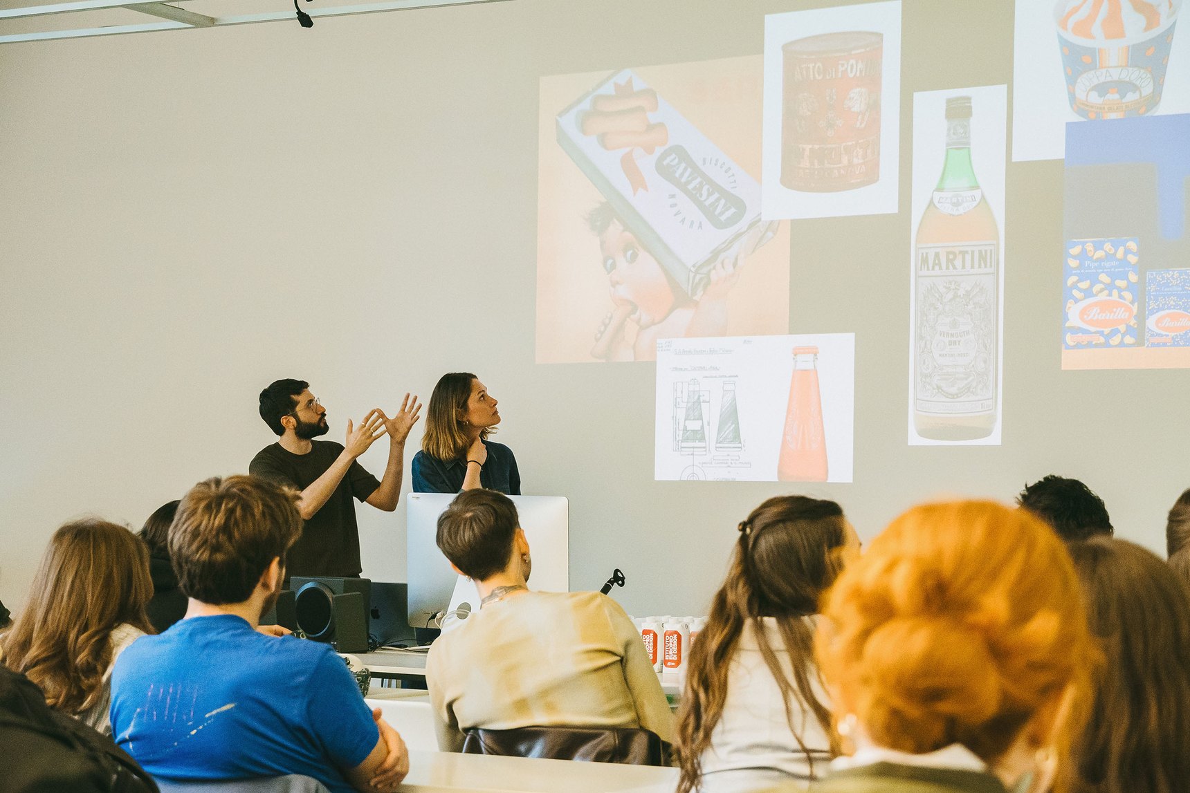 Pubblico seduto in un'aula davanti a due relatori, con manifesti e illustrazioni proiettati sulla parete