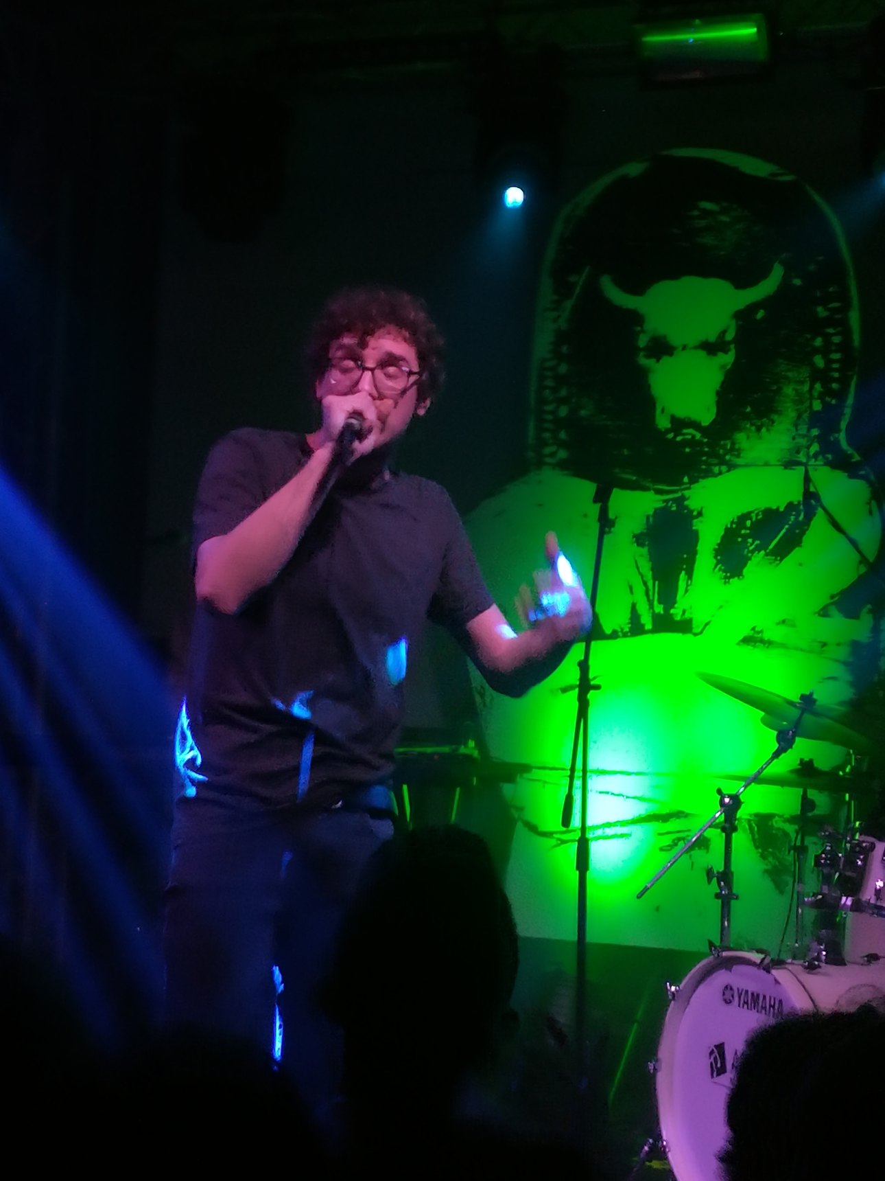 Willie Peyote singing into the microphone on a stage lit in green and blue, with a drum kit behind him