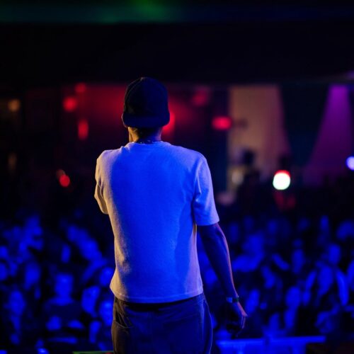 Un artista di spalle, con cappellino e maglietta bianca, sul palco davanti al pubblico nelle luci blu di un concerto