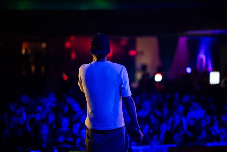 Un artista di spalle, con cappellino e maglietta bianca, sul palco davanti al pubblico nelle luci blu di un concerto