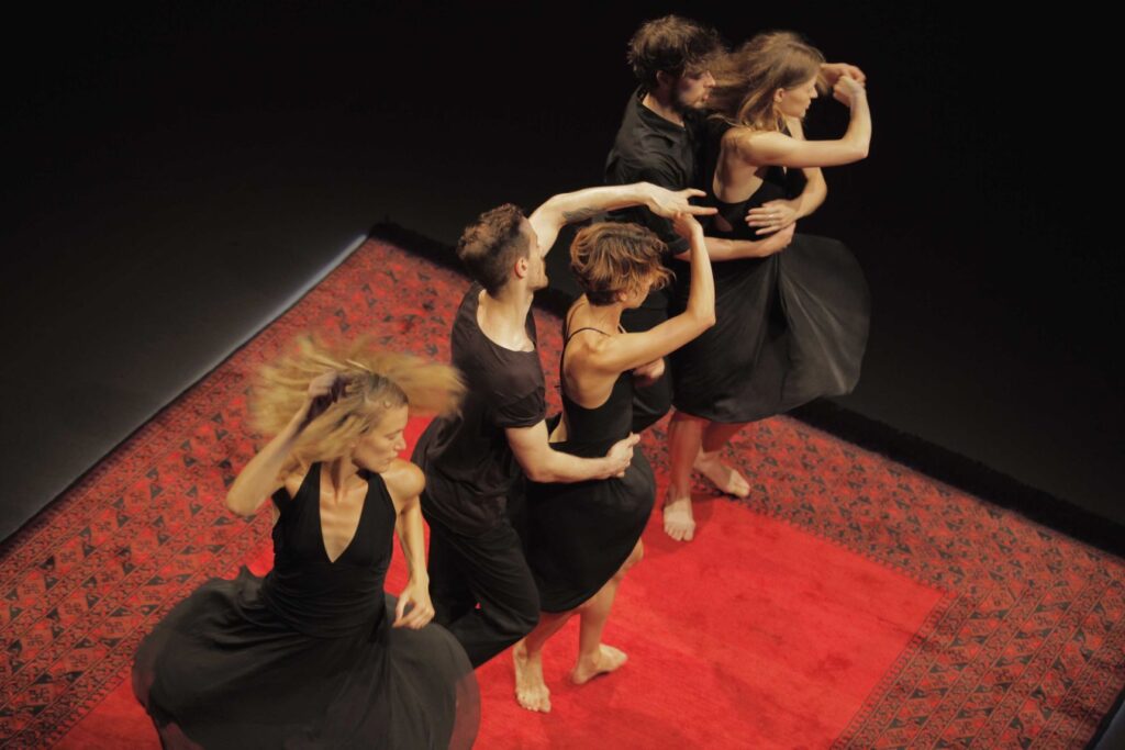 BACH to SACRE di Emanuel Gat Dance: danzatrici in abiti neri con le braccia sopra la testa, su un tappeto rosso