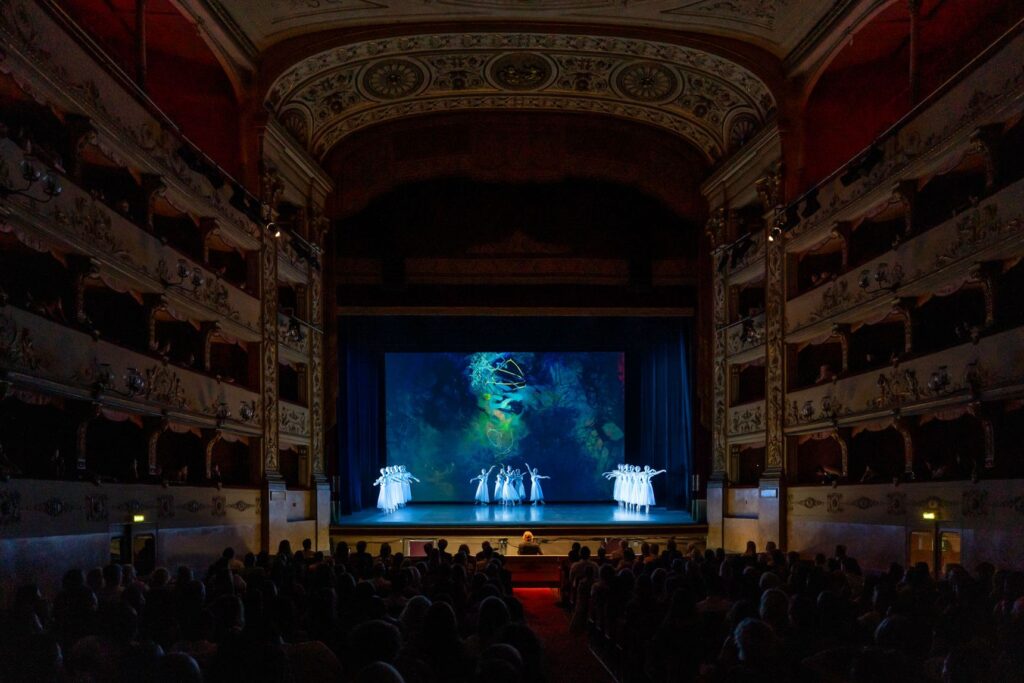 Il balletto in scena al New Generation Festival 2025, visto dal fondo della platea di un teatro con i palchi illuminati