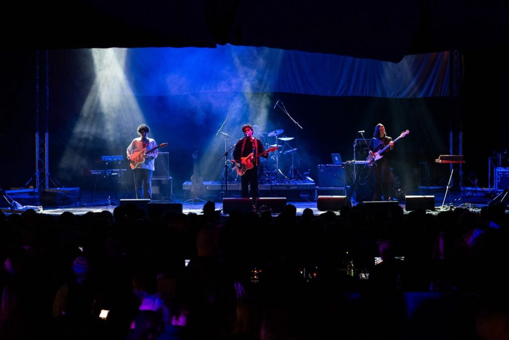 Three musicians with guitars and bass on a stage bathed in blue light, with spotlight beams from above and the audience in shadow
