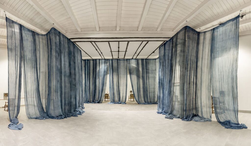 Veduta della mostra all'ex Teatro dell'Oriuolo: lunghe tende di garza blu scendono dal soffitto bianco e dividono la sala